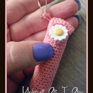 May include: A pink crocheted item with a white daisy embellishment and a silver-colored clip. The text "Unique As I Am" is at the bottom. The item is held in a hand with a purple nail.