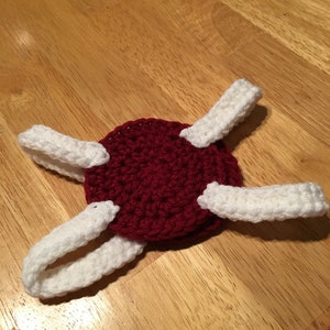 Puede incluir: Un art&iacute;culo hecho a mano con ganchillo con una base circular de color rojo oscuro con cuatro acentos blancos, curvos y rectangulares. El art&iacute;culo est&aacute; colocado sobre una superficie de madera. El ganchillo parece estar cosido con fuerza.