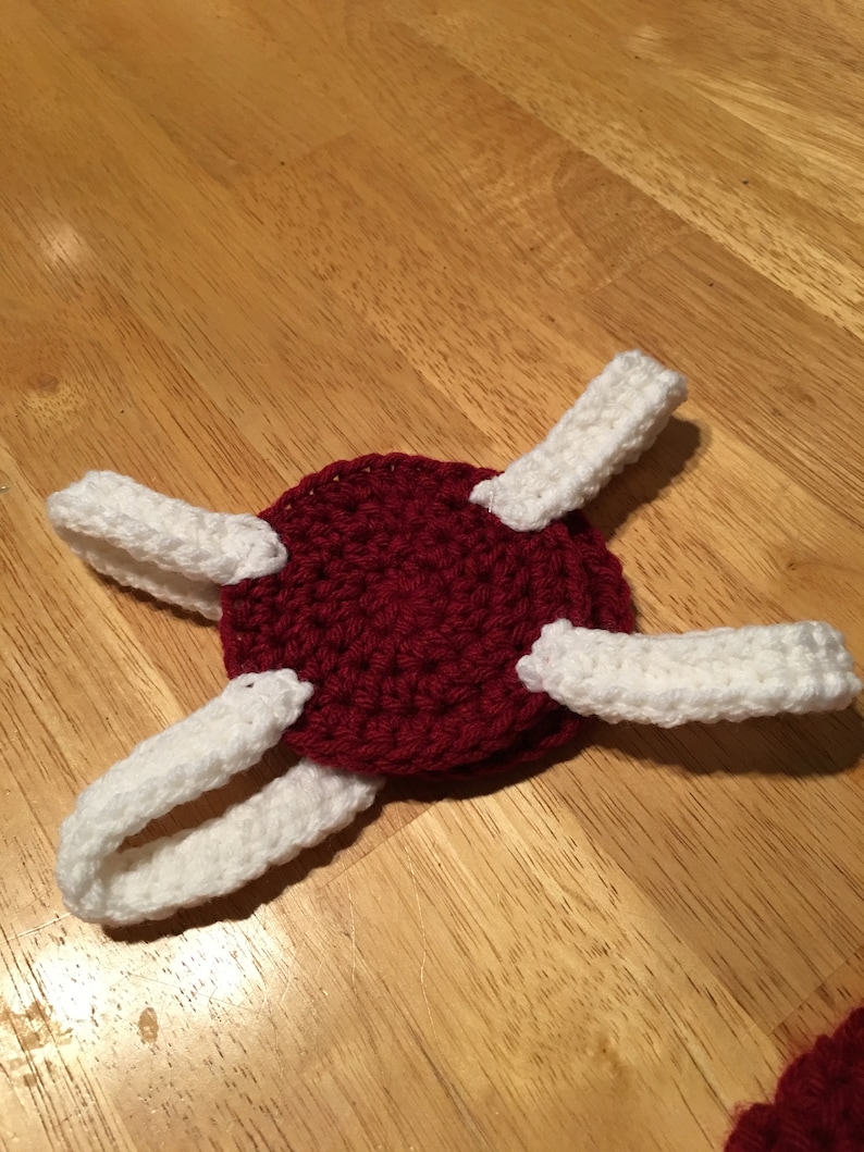 Puede incluir: Un art&iacute;culo hecho a mano con ganchillo con una base circular de color rojo oscuro con cuatro acentos blancos, curvos y rectangulares. El art&iacute;culo est&aacute; colocado sobre una superficie de madera. El ganchillo parece estar cosido con fuerza.