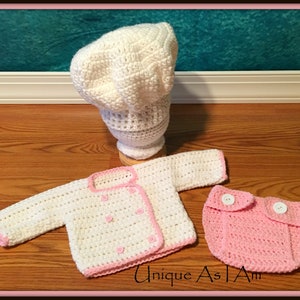 May include: A white crocheted chef hat, a white crocheted jacket with pink trim and two white buttons, and a pink crocheted diaper with two white buttons. The text "Unique As I Am" is visible in the bottom right corner of the image.