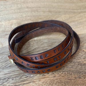 May include: A brown leather wrap bracelet featuring a metallic snap closure. The leather strap is embossed with the text KEEP THE BLOOD AT MY HEART I HAVE BEEN.