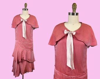 May include: A vintage rose-colored dress with a matching cape-like collar and a cream-colored bow. The dress features a tiered skirt and is displayed on a mannequin against a pink background. The dress is a vintage style.
