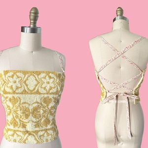 May include: A sleeveless crop top made from yellow and white floral patterned terry cloth fabric. The front features a straight neckline, while the back displays a crisscross lace-up design with thin straps featuring a small red and white floral print.