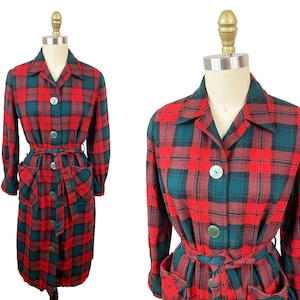 Op de afbeelding: Een rood-groene geruite jurk met lange mouwen, een kraag en een riem. De jurk heeft twee zakken aan de voorkant.