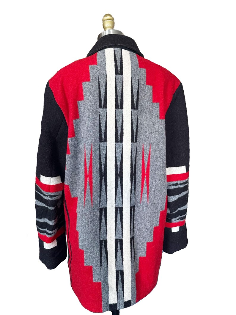 May include: A wool coat with a geometric pattern in red, gray, black, and white. The coat features a black collar and sleeves with red and white accents. The back of the coat showcases a striking diamond-shaped design.