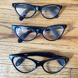 May include: Three pairs of black cat-eye glasses with clear lenses. The glasses are arranged in a vertical stack, with the smallest pair at the top and the largest pair at the bottom. The glasses are made in Italy.