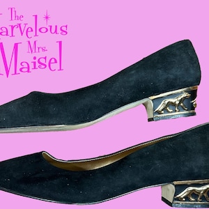 May include: A pair of black suede pumps with a low block heel featuring a gold-toned metal panther accent on the side. The text The Marvelous Mrs. Maisel is printed in bright pink in the upper left corner of the image.