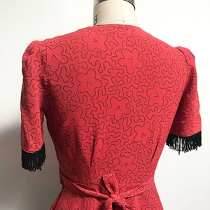 Pode incluir: Uma blusa vermelha com um padr&atilde;o abstrato preto e acabamento em franjas pretas. A blusa tem um la&ccedil;o na cintura.