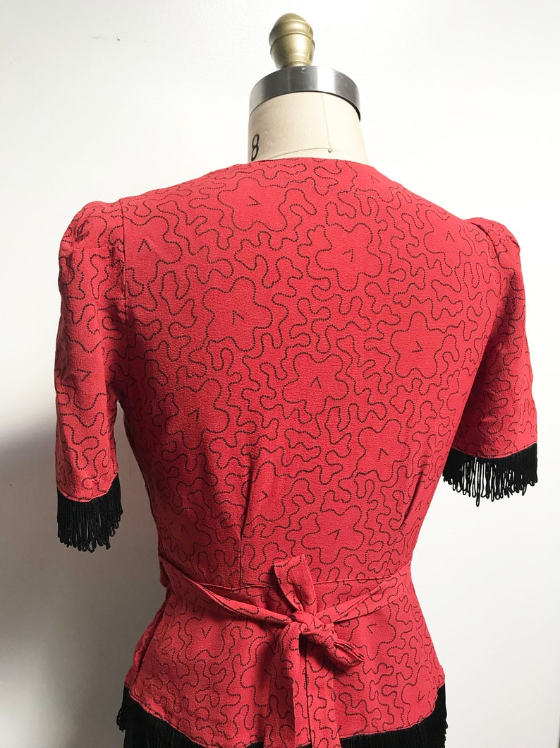 Pode incluir: Uma blusa vermelha com um padr&atilde;o abstrato preto e acabamento em franjas pretas. A blusa tem um la&ccedil;o na cintura.
