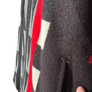 May include: Close-up of a dark brown wool garment with a geometric pattern. The design includes white and gray shapes, with a bold red stripe down the center. The garment has a pocket.