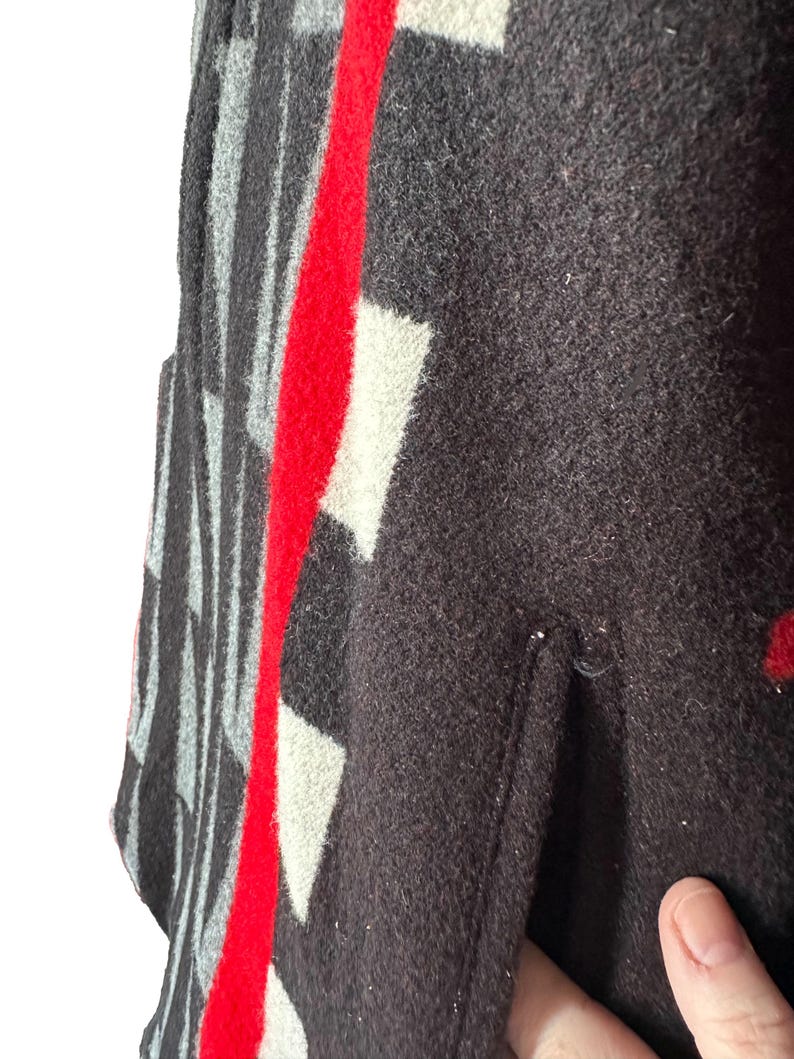May include: Close-up of a dark brown wool garment with a geometric pattern. The design includes white and gray shapes, with a bold red stripe down the center. The garment has a pocket.
