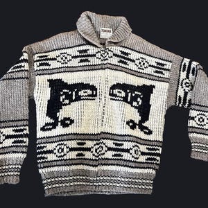 May include: A gray, white, and black knitted sweater with a shawl collar. The sweater features a geometric pattern and a central design with the text "13". The label reads "TUNDRA".