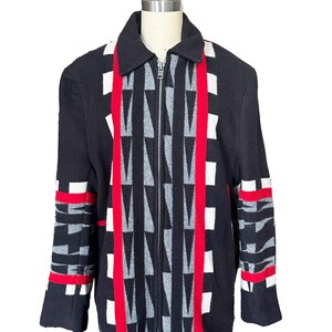 May include: Black zip-up jacket with a geometric pattern. The jacket features a central design of gray and white triangles, red vertical stripes, and white and red accents on the sleeves. The jacket is displayed on a mannequin.