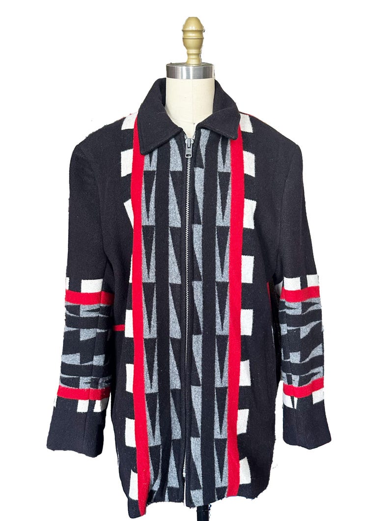 May include: Black zip-up jacket with a geometric pattern. The jacket features a central design of gray and white triangles, red vertical stripes, and white and red accents on the sleeves. The jacket is displayed on a mannequin.