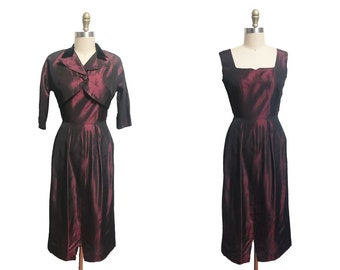 May include: A burgundy taffeta dress with a matching bolero jacket. The dress has a fitted bodice and a full skirt. The bolero jacket has a shawl collar and three-quarter sleeves.