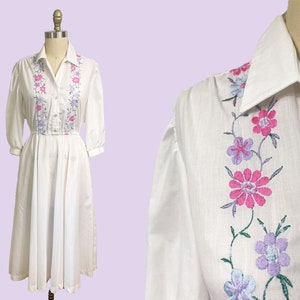 May include: White cotton dress with embroidered pink and purple flowers. The dress has a collared neckline and 3/4 sleeves.