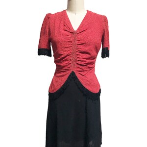 Pode incluir: Um vestido vermelho com saia preta e acabamento de franjas pretas. O vestido tem um corpete justo e uma cintura franzida. A saia &eacute; cheia e fluida.