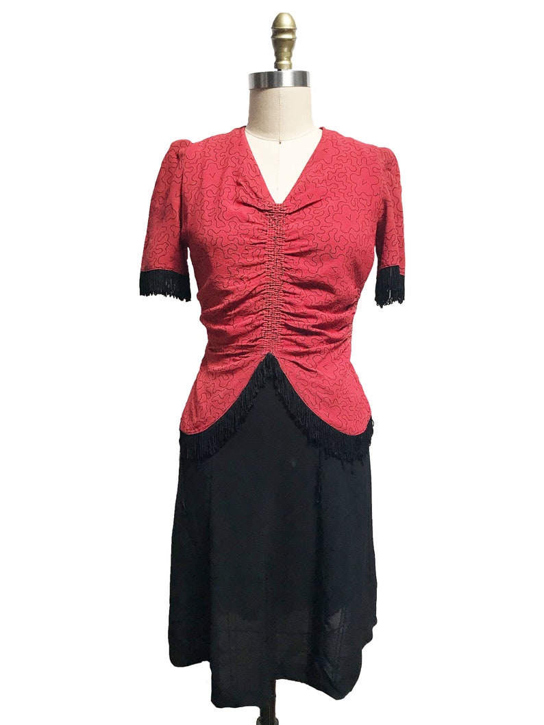 Pode incluir: Um vestido vermelho com saia preta e acabamento de franjas pretas. O vestido tem um corpete justo e uma cintura franzida. A saia &eacute; cheia e fluida.