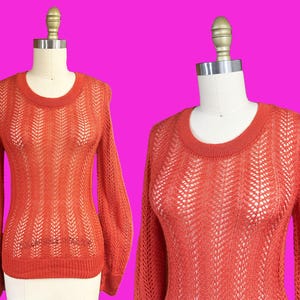 Puede incluir: Un suéter calado de color naranja quemado con cuello redondo y mangas largas. El patrón de punto presenta un diseño de chevrón vertical. El suéter se exhibe en un maniquí sobre un fondo rosa brillante.