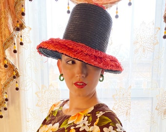 May include: A woman wearing a black and pink straw hat with a wide brim and green earrings. She is wearing a brown floral print dress.