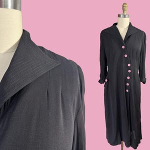 Könnte beinhalten: Ein schwarzes Kleid mit langen Ärmeln und Kragen. Das Kleid hat rosa Knöpfe auf der Vorderseite und an den Manschetten. Das Kleid wird auf einer Schaufensterpuppe vor rosa Hintergrund präsentiert.