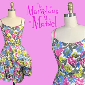 May include: A vintage floral print dress with a bubble skirt, displayed on a mannequin. The dress features a colorful pattern of pink, blue, and yellow flowers on a white background. It has thin pink straps and a matching belt. The text "The Marvelous Mrs. Maisel" is in the background.