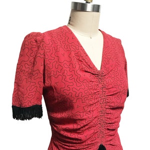Pode incluir: Um top vermelho com um padr&atilde;o abstrato preto e branco. O top tem um decote em V e mangas curtas com acabamento em franjas pretas.