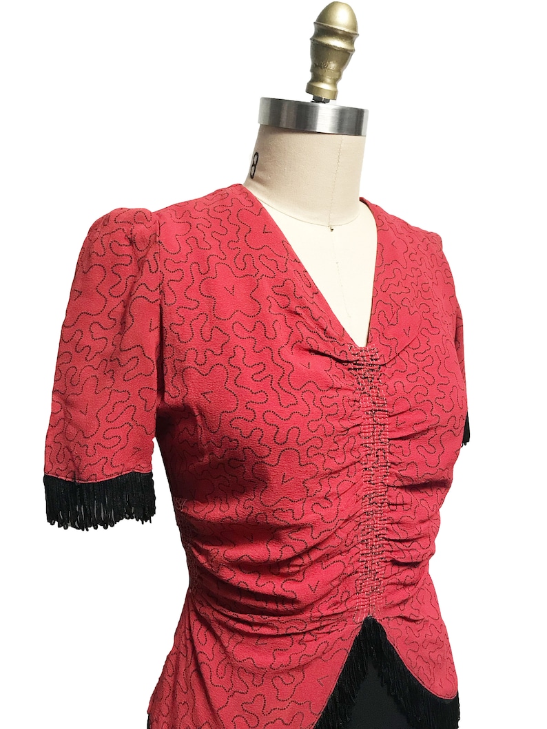 Pode incluir: Um top vermelho com um padr&atilde;o abstrato preto e branco. O top tem um decote em V e mangas curtas com acabamento em franjas pretas.
