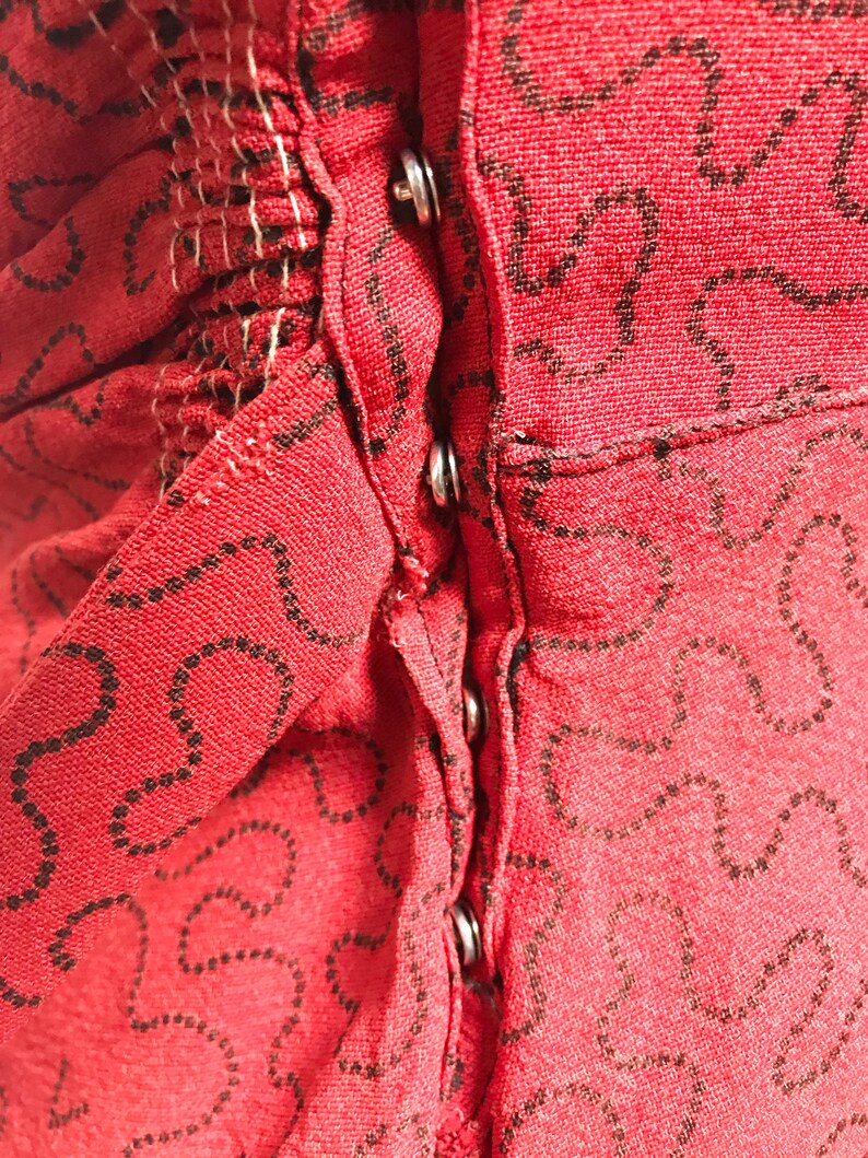 Pode incluir: Close-up de um tecido vermelho com um padr&atilde;o preto. O tecido tem uma fileira de bot&otilde;es prateados no centro.