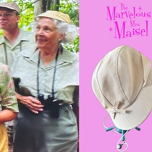 May include: A beige fabric hat with a structured crown and a short, curved brim featuring a small decorative bow detail on the side. The hat is displayed on a stand against a pink background with the text The Marvelous Mrs. Maisel.