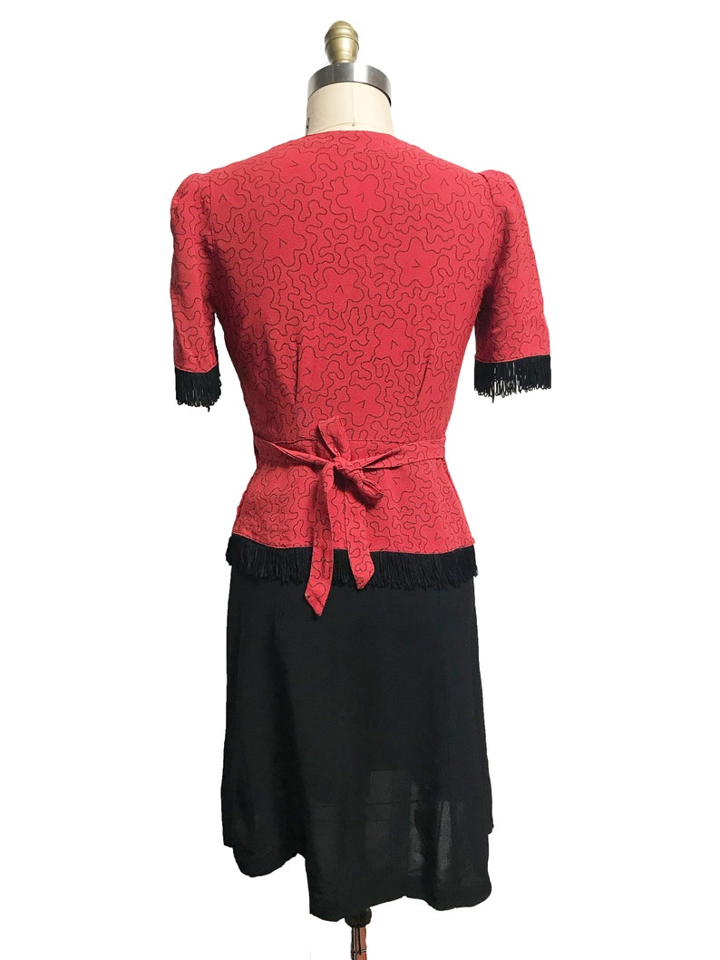 Pode incluir: Um vestido vermelho com uma borda de franjas pretas e uma saia preta. O vestido tem um decote em V e um la&ccedil;o na cintura.