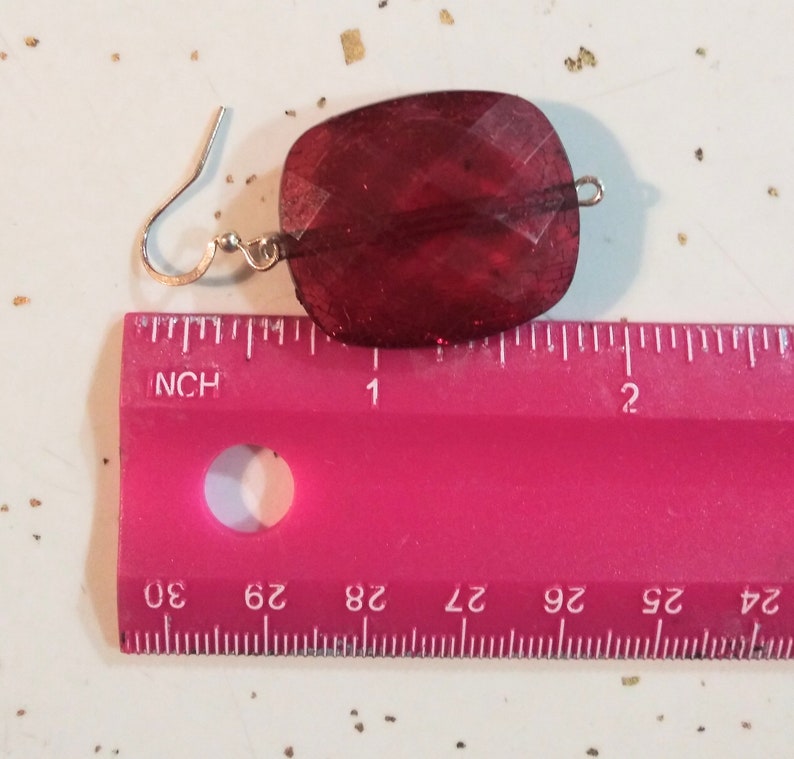 Pode incluir: Um par de brincos de vidro vermelho com um gancho de prata. Os brincos medem aproximadamente 2,5 cm de comprimento.