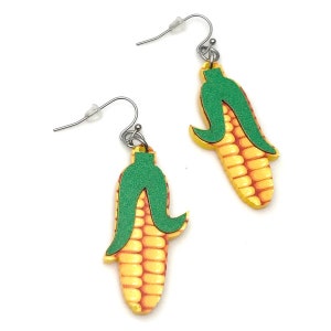 May include: A pair of dangle earrings featuring a corn on the cob design. The earrings are made of wood and painted yellow, green, and red. The corn is depicted with a green husk and yellow kernels.