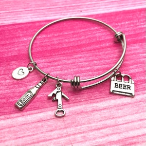 May include: A silver charm bracelet with a beer bottle, bottle opener, heart, and a six-pack of beer charm. The bracelet is on a pink background.