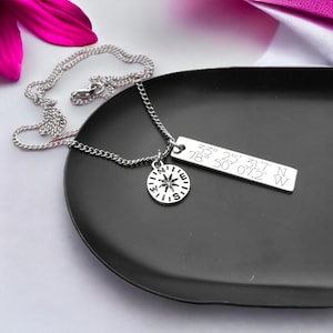 Puede incluir: Un collar de plata con un dije de br&uacute;jula y una etiqueta rectangular con las coordenadas "33&deg; 25' 31.7" N 78&deg; 50' 07.2" W" grabadas en ella.