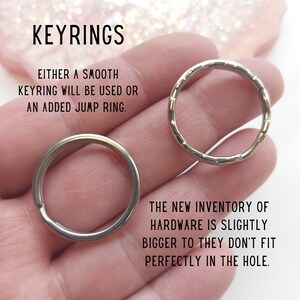 May include: Two silver keyrings, one smooth and one with a textured edge, are held in a hand. The text "KEYRINGS" is at the top of the image. The text "EITHER A SMOOTH KEYRING WILL BE USED OR AN ADDED JUMP RING." is below the keyrings. The text "THE NEW INVENTORY OF HARDWARE IS SLIGHTLY BIGGER TO THEY DON'T FIT PERFECTLY IN THE HOLE." is at the bottom of the image.