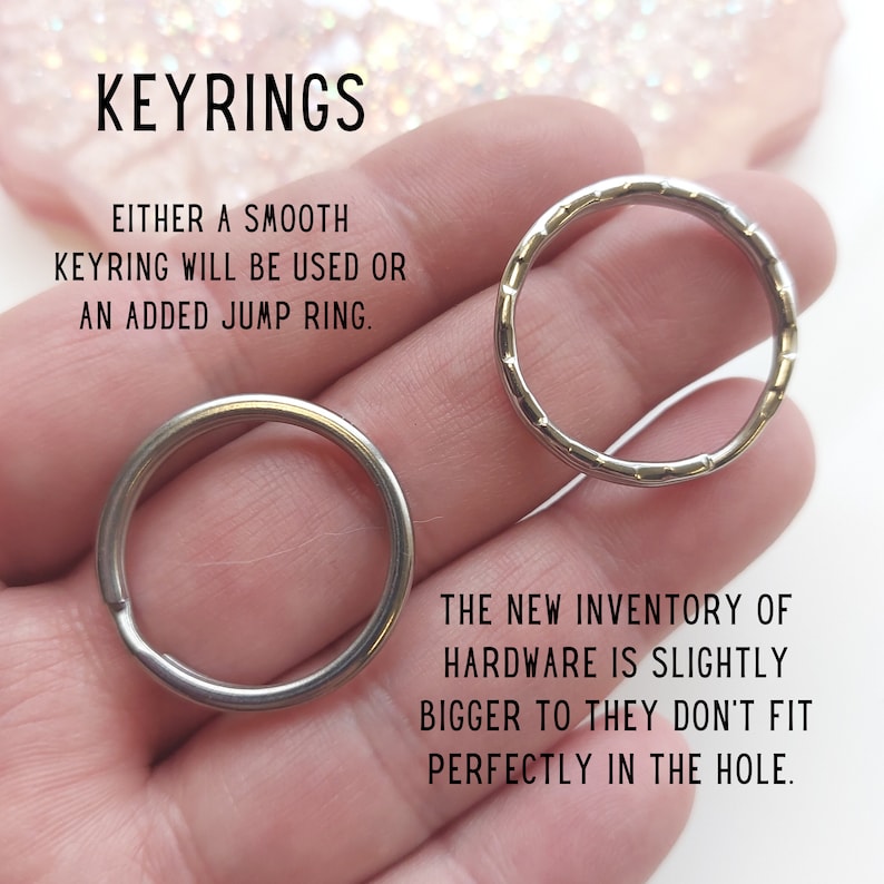 May include: Two silver keyrings, one smooth and one with a textured edge, are held in a hand. The text "KEYRINGS" is at the top of the image. The text "EITHER A SMOOTH KEYRING WILL BE USED OR AN ADDED JUMP RING." is below the keyrings. The text "THE NEW INVENTORY OF HARDWARE IS SLIGHTLY BIGGER TO THEY DON'T FIT PERFECTLY IN THE HOLE." is at the bottom of the image.