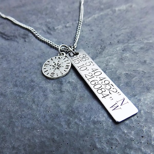 Puede incluir: Collar de plata con un dije de br&uacute;jula y un dije rectangular grabado con coordenadas: "36.5404952", "N" y "115.10216084", "W".