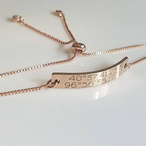 May include: A rose gold adjustable bracelet with a rectangular charm engraved with the coordinates 40°37'41.25"N 96°56'35.17"W.