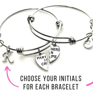 May include: Two silver bangle bracelets with charms. One charm says "Part in Crime" and the other says "Partners in Crime". The bracelets have a heart charm, a handcuff charm, and a letter charm. Choose your initials for each bracelet.