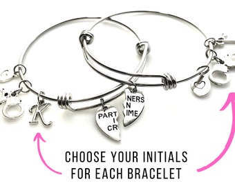 May include: Two silver bangle bracelets with charms. One charm says "Part in Crime" and the other says "Partners in Crime". The bracelets have a heart charm, a handcuff charm, and a letter charm. Choose your initials for each bracelet.