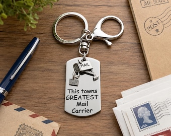 Könnte beinhalten: Ein silberfarbener Metallschlüsselanhänger mit einem rechteckigen Anhänger, der mit dem Text 'This towns GREATEST Mail Carrier' graviert ist. Daran befestigt ist ein kleiner Briefkasten-Anhänger mit der Aufschrift 'MAIL' sowie ein kleines Paket.