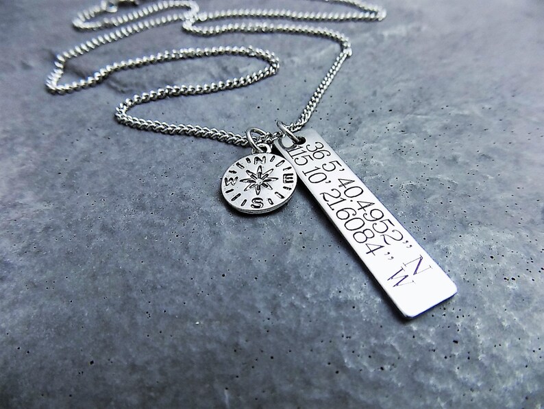 Puede incluir: Un collar de plata con un dije de br&uacute;jula y un dije rectangular con las coordenadas "36.54049523, N 115.10216084, W" grabadas en &eacute;l.