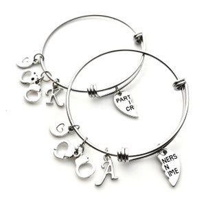 May include: Two silver bangle bracelets with charms. One bracelet has a heart charm that says "PART I CR" and the other bracelet has a heart charm that says "NERS N IME", a handcuff charm, and a letter "A" charm.