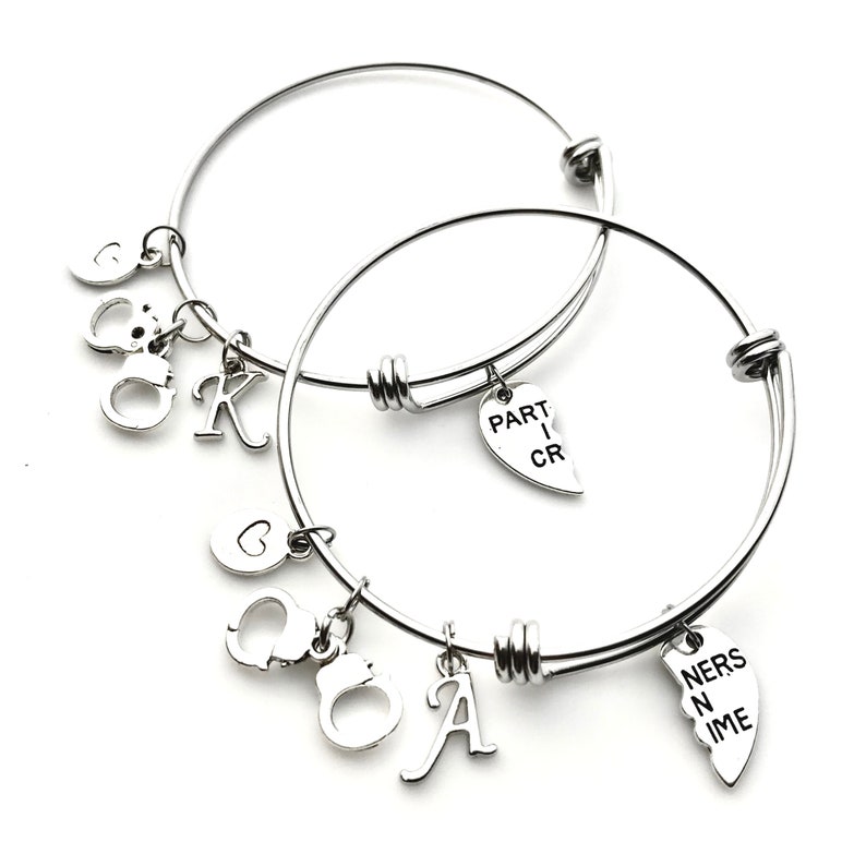 May include: Two silver bangle bracelets with charms. One bracelet has a heart charm that says "PART I CR" and the other bracelet has a heart charm that says "NERS N IME", a handcuff charm, and a letter "A" charm.