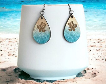 May include: A pair of teardrop-shaped wooden earrings with a sea turtle design. The earrings are painted with a gradient of blue and brown, resembling a beach scene.