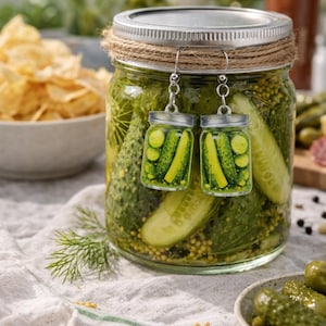 K&ouml;nnte beinhalten: Ohrringe zum Aufh&auml;ngen in Form kleiner Einmachgl&auml;ser mit Essiggurken, die vor einem gro&szlig;en Einmachglas mit Essiggurken h&auml;ngen. Die Ohrringe haben silberfarbene Deckel und klare Gl&auml;ser, die mit gr&uuml;nen Essiggurken gef&uuml;llt sind. Im Hintergrund ist eine Sch&uuml;ssel mit Chips zu sehen.