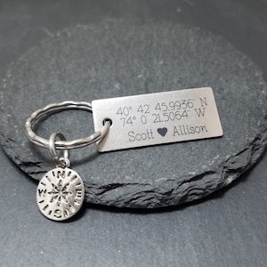 May include: A silver keychain with a compass charm and a rectangular tag engraved with the coordinates 40&deg; 42 45.9936" N 74&deg; 0&prime; 21.5064" W and the names Scott and Allison.