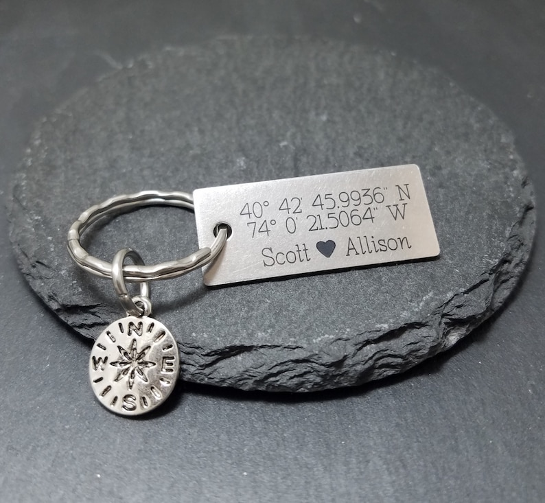May include: A silver keychain with a compass charm and a rectangular tag engraved with the coordinates 40&deg; 42 45.9936" N 74&deg; 0&prime; 21.5064" W and the names Scott and Allison.