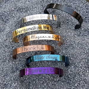 May include: A set of seven metal bracelets with different engraved text. The bracelets are in various colors, including silver, gold, copper, blue, and purple. The text on the bracelets includes "My favorite fine series", "Personalize Me", "Megan Kyle", "Thankful", "Bold Script", and "Typewriter".